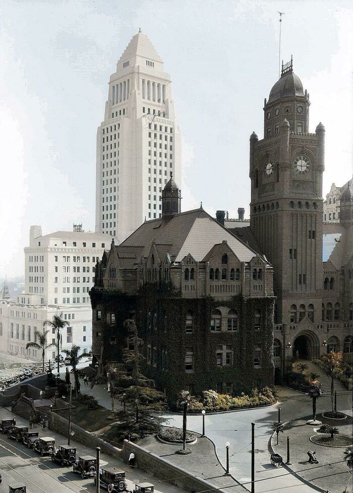 A historical photo showing a tall building behind a vintage building covered in ivy, with old cars parked on the street.