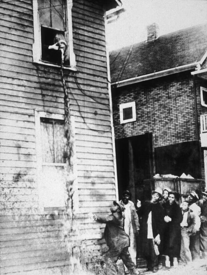 A man rescues a cat from a burning building using a rope while people watch from below, a moment captured in historical photos.