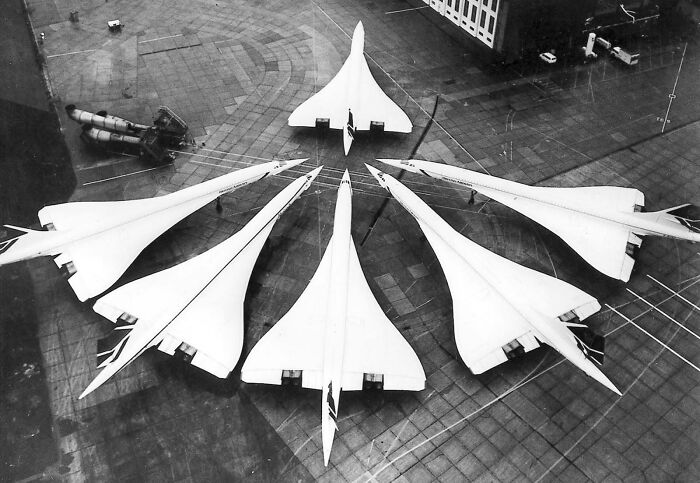 A high-angle view of several Concorde supersonic jets arranged in a semi-circle on an airfield, showing historical photos.