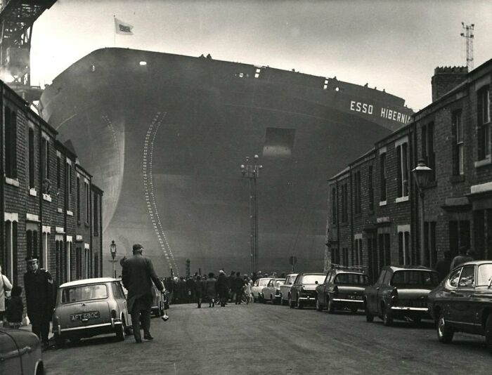 Historical photo of a huge ship, Esso Hibernia, dwarfing a street with parked cars and onlookers, a vivid past scene.