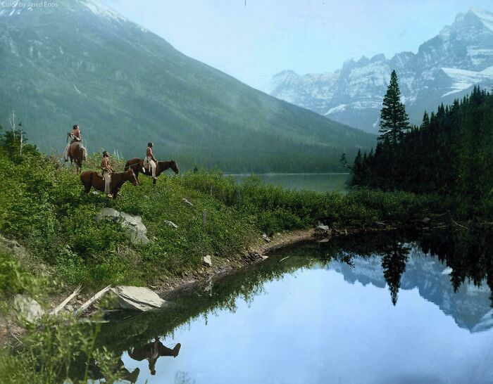 A historical photo of people on horseback by a serene lake, surrounded by mountains, revealing a beautiful past landscape.