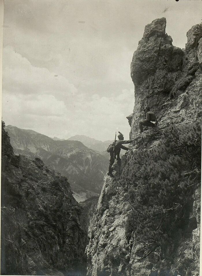 A striking historical photo captures two soldiers scaling a steep, rocky mountain face amidst a vast, rugged landscape.
