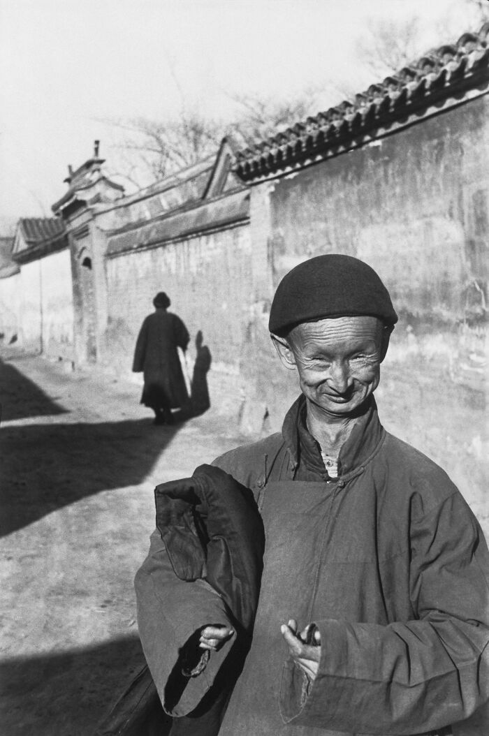 A historical photo features an elderly smiling man in traditional attire standing on a street, with another person in the background.