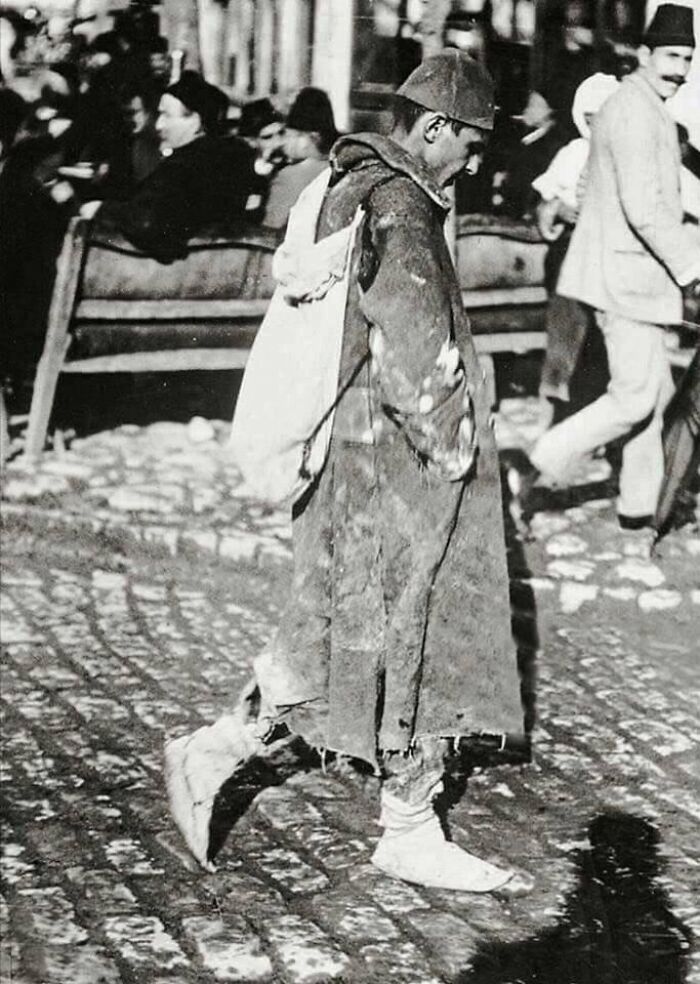 A man in worn clothing and a cap walks down a cobblestone street, carrying a sack, in a historical photo of a past era.