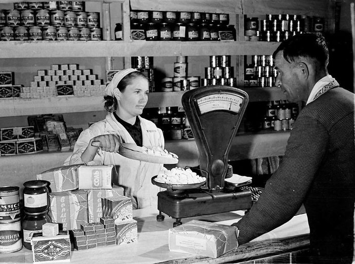 A female store clerk uses a scale to weigh items for a male customer in a historical photo of a past shop.
