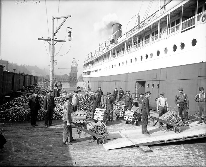Workers load crates onto a ship from a dockside with many historical photos and a past industrial scene.