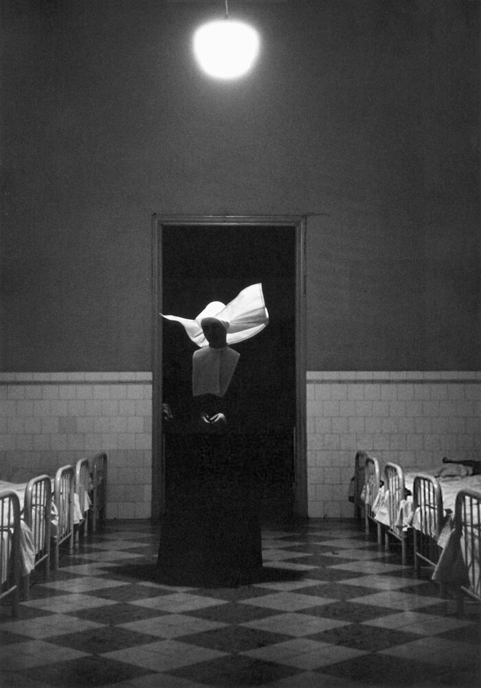 A nun in a habit stands in a dimly lit hallway between rows of beds in a historical photo of a past hospital.