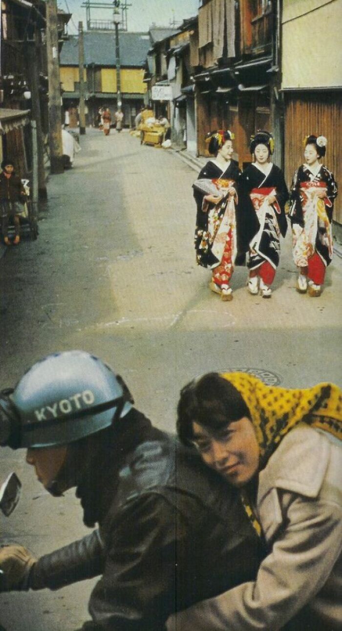 A historical photo of a street in Kyoto with a motorcycle rider and passengers, and three women in kimonos walking, a past scene.