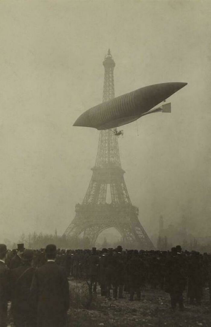 A historical photo of a dirigible flying above the Eiffel Tower with a crowd below, remembering a past event.