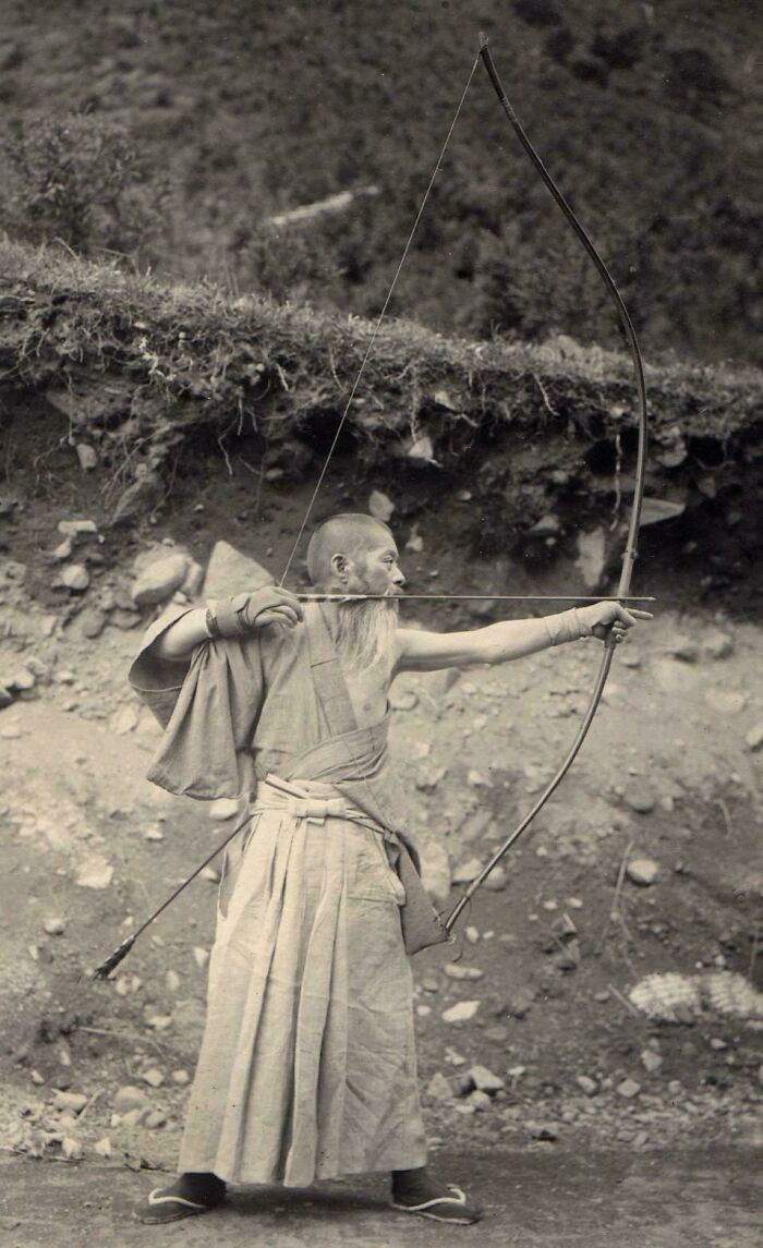 Historical photo of a man in traditional attire aiming a bow and arrow, capturing a past moment.