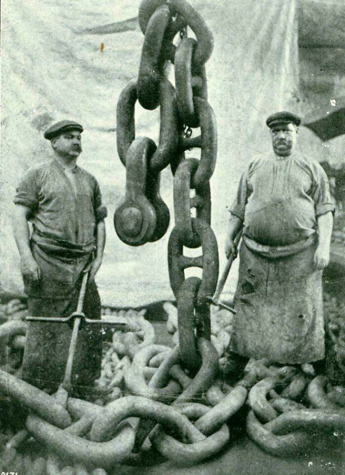 Two strong men standing beside a colossal anchor chain, highlighting a fascinating historical photo.