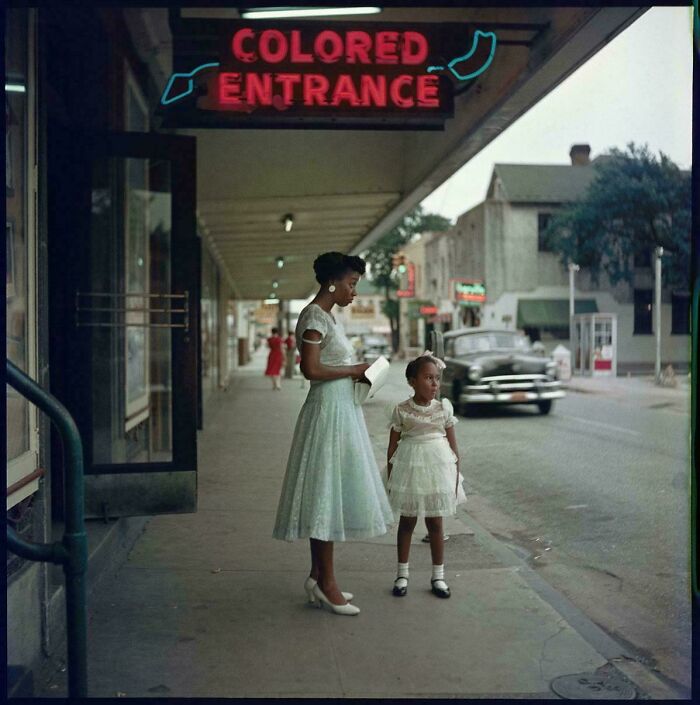 Historical photo showing a woman and a young girl standing on a street, with a neon sign for a segregated entrance.