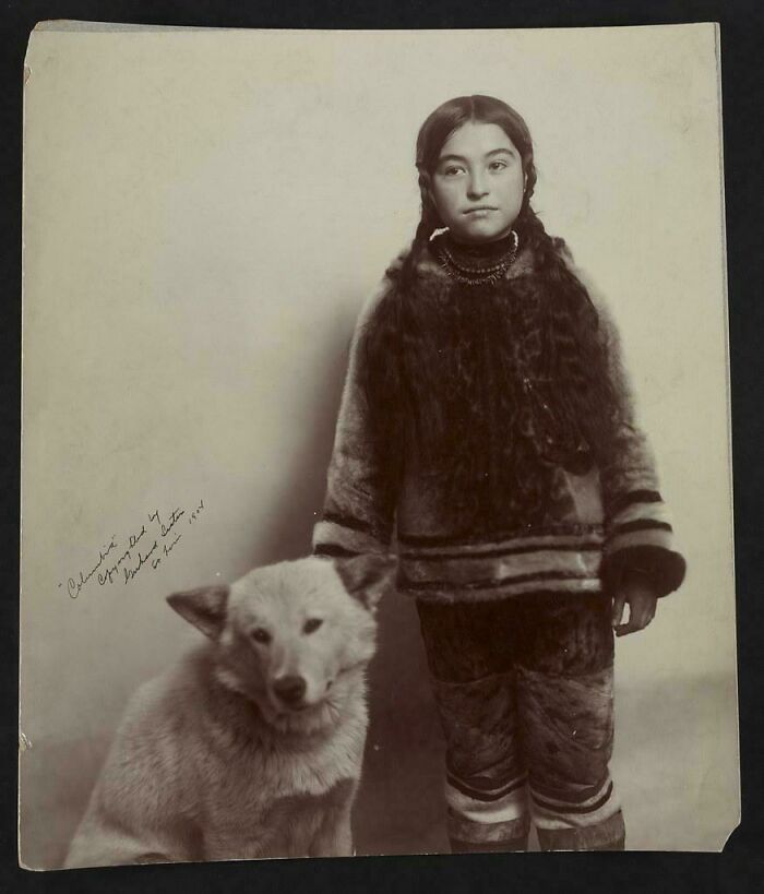 Historical photo of a young indigenous girl in traditional fur clothing standing next to a white dog.