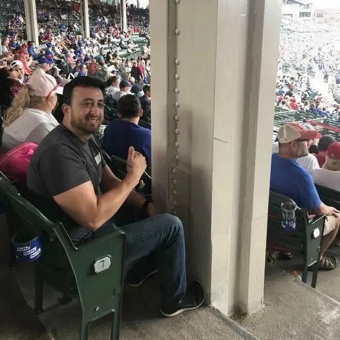 A construction fail: a man giving a thumbs up from a stadium seat directly behind a large pillar.