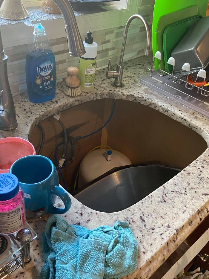 A kitchen sink with the basin removed, exposing plumbing and a water filter tank, showing a problem needing help fixing.