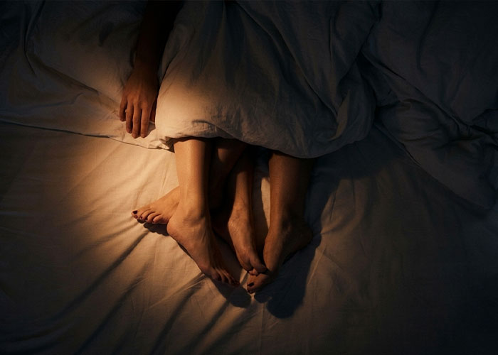 A couple's feet intertwined under a blanket, illustrating the intimacy of the Queening position. A couple's feet intertwined under a blanket, illustrating the intimacy of the Queening position.