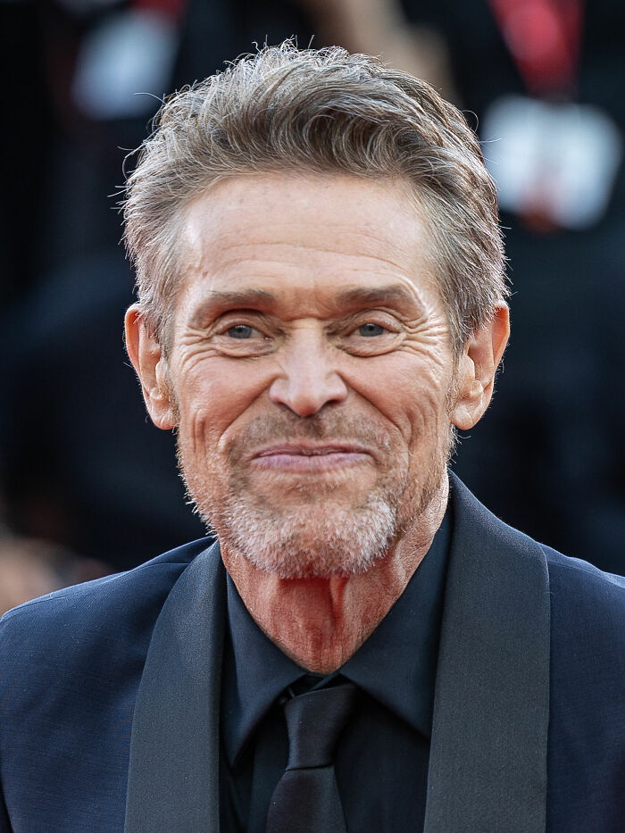 Close-up of Willem Dafoe smiling, showcasing his bio and career highlights with a grey beard and dark suit.