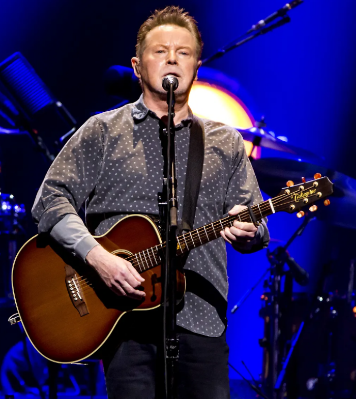 Don Henley performing live, singing into a microphone and playing an acoustic guitar, showcasing his iconic career highlights.