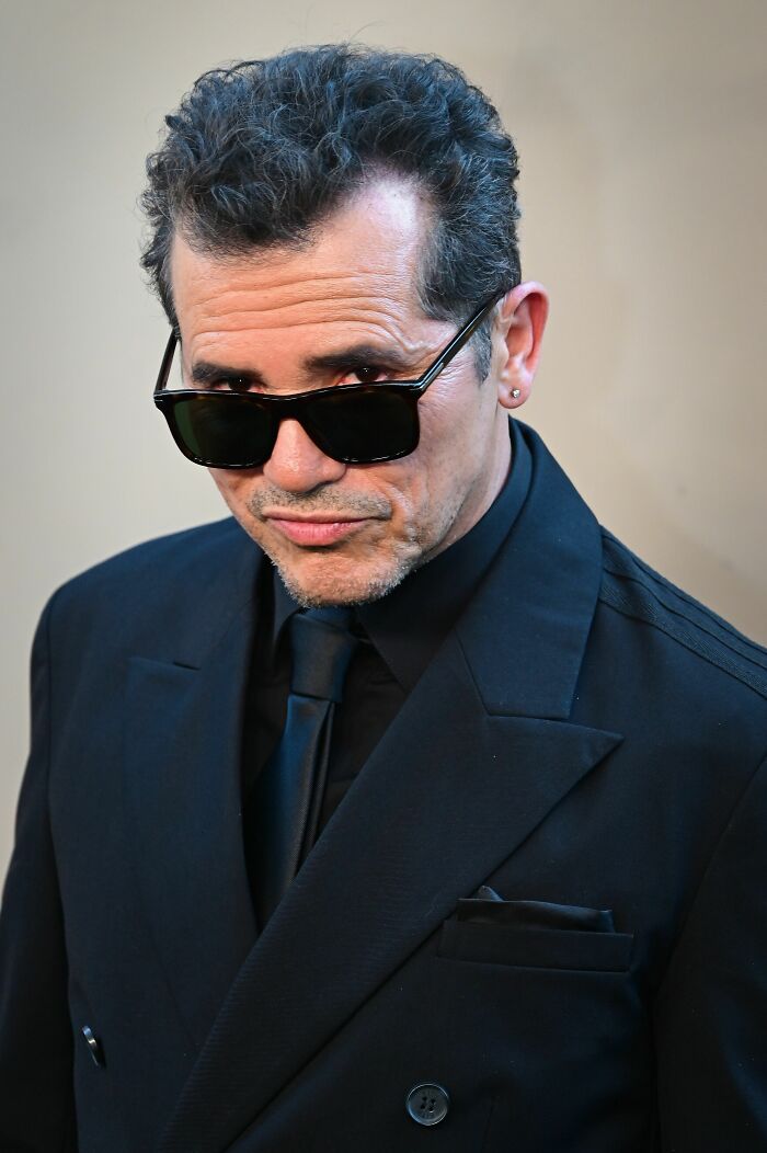 John Leguizamo, actor, wearing sunglasses and a black suit, looking directly at the camera with a slight smirk.