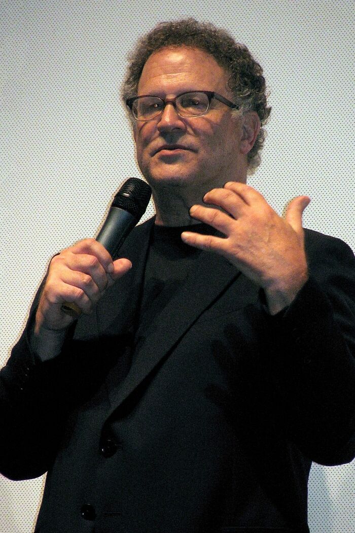 Albert Brooks giving a presentation, holding a microphone, showcasing his career highlights and bio.