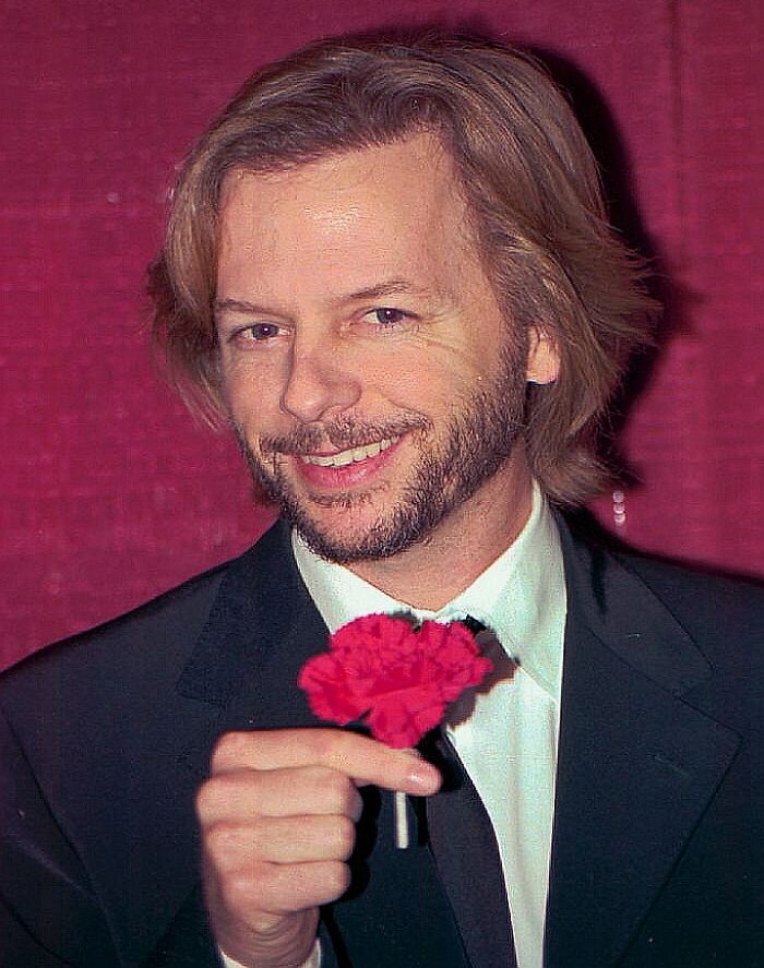 David Spade, actor and comedian, smiles while holding a red flower. This image highlights David Spade's career.