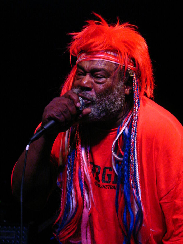 George Clinton performs live, with red hair and colorful braids, singing into a microphone on stage.