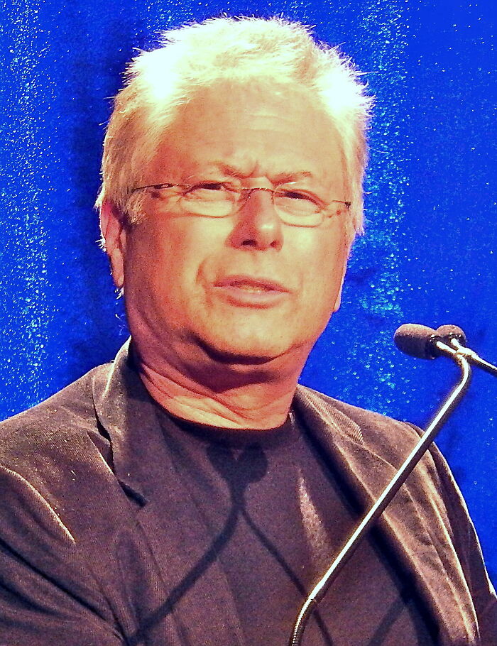 Alan Menken with white hair and glasses, speaking into a microphone, highlighting his bio and career.