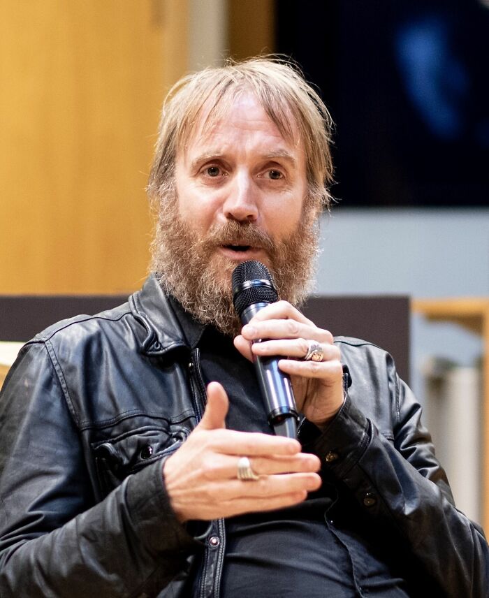 Rhys Ifans speaks into a microphone, showcasing career highlights with his distinctive look, wearing a black leather jacket.