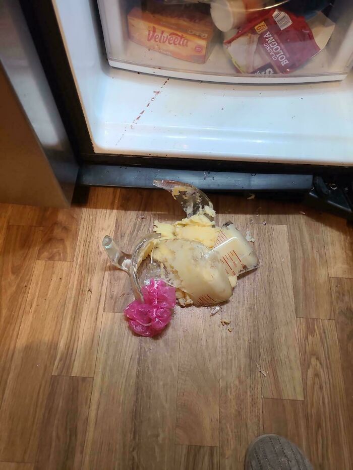 A broken glass measuring cup with spilled potato salad on a wooden floor, creating a worse day mess.
