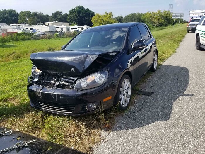 A black car with significant front-end damage on the side of a road, part of 87 People Who Are Having A Way Worse Day Than You.