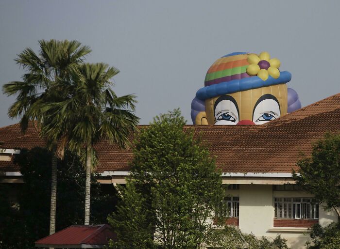 A large clown-faced hot air balloon peeking over a building, having a worse day than you.