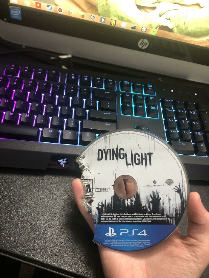 A hand holding a shattered Dying Light PS4 game disc in front of a colorful gaming keyboard, illustrating a truly bad day.