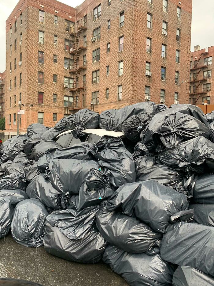 A massive pile of black trash bags filling a street, almost completely covering a car, with apartment buildings in the background.