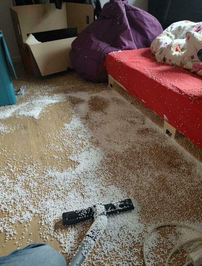 A person having a worse day, looking at a floor covered in white foam pellets near a bed and a vacuum.