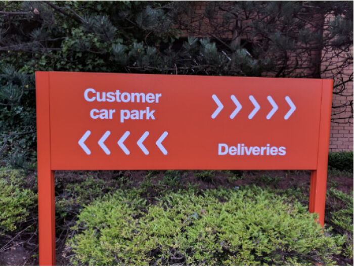 A red sign with arrows pointing to the right for customer car park and to the left for deliveries, hilariously failed at clear direction.