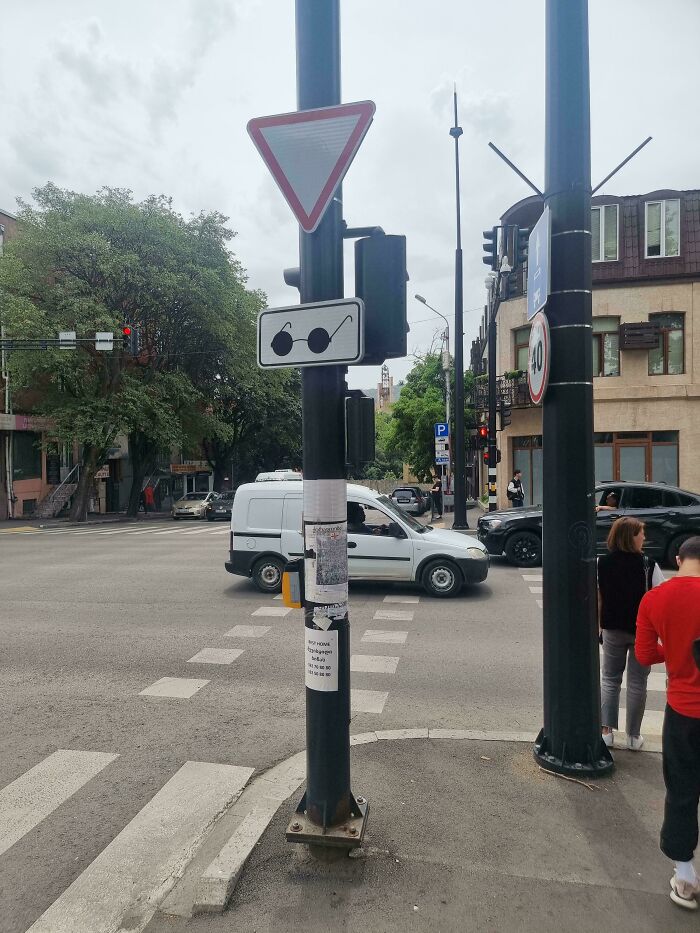 A street scene with a hilariously failed sign of eyeglasses on a pole, overlooking a busy intersection with cars and pedestrians.