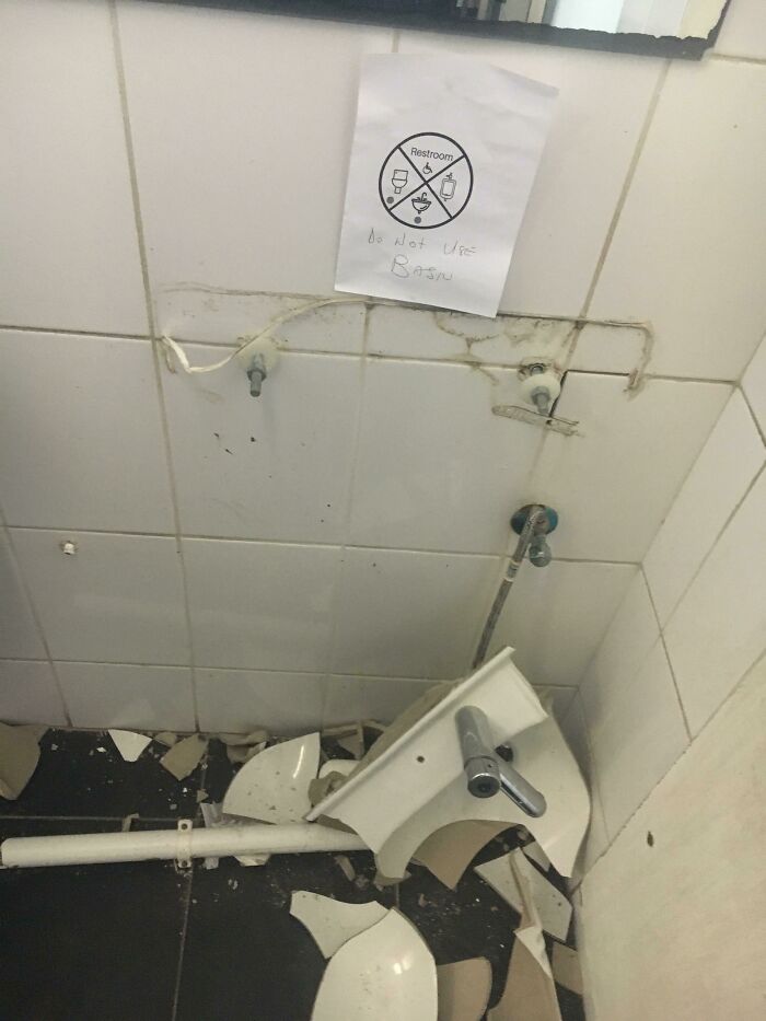 A bathroom sign that hilariously failed, taped above a completely shattered basin, stating 'Do Not Use Basin'.
