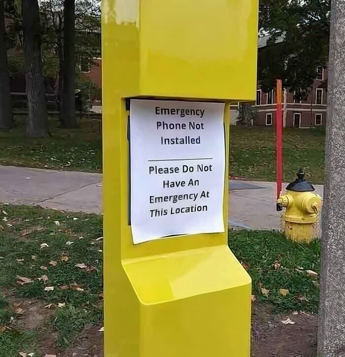 A hilariously failed sign on a yellow emergency phone post reads, Emergency Phone Not Installed. Please Do Not Have An Emergency At This Location.