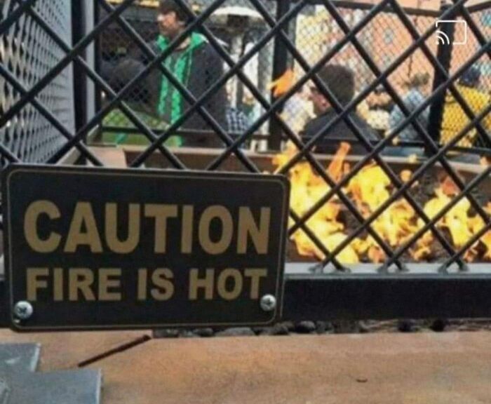 A hilariously failed sign reading CAUTION FIRE IS HOT next to an actual burning fire pit.