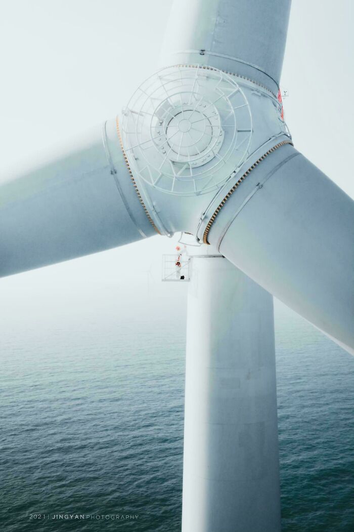 A worker standing on a platform on a colossal wind turbine blade over the ocean, revealing the true scale of the structure.