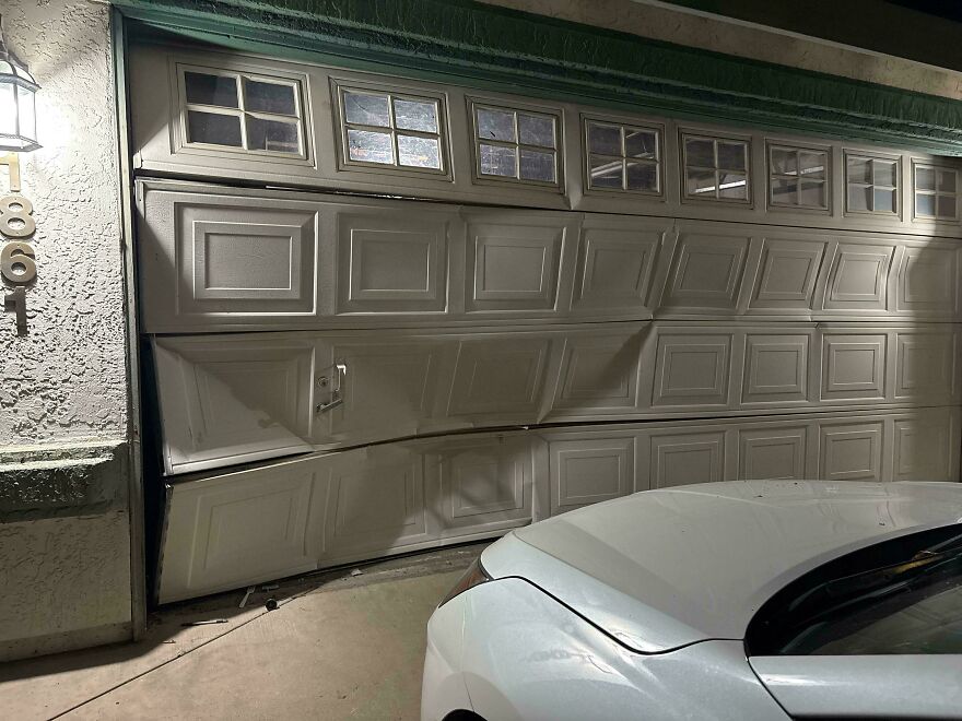 A severely bent and damaged garage door with a white car in front, showing an expensive accident.