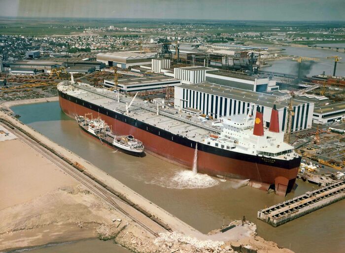 An aerial view of a massive ship dwarfing a smaller vessel at a shipyard, illustrating true scale in shipbuilding.