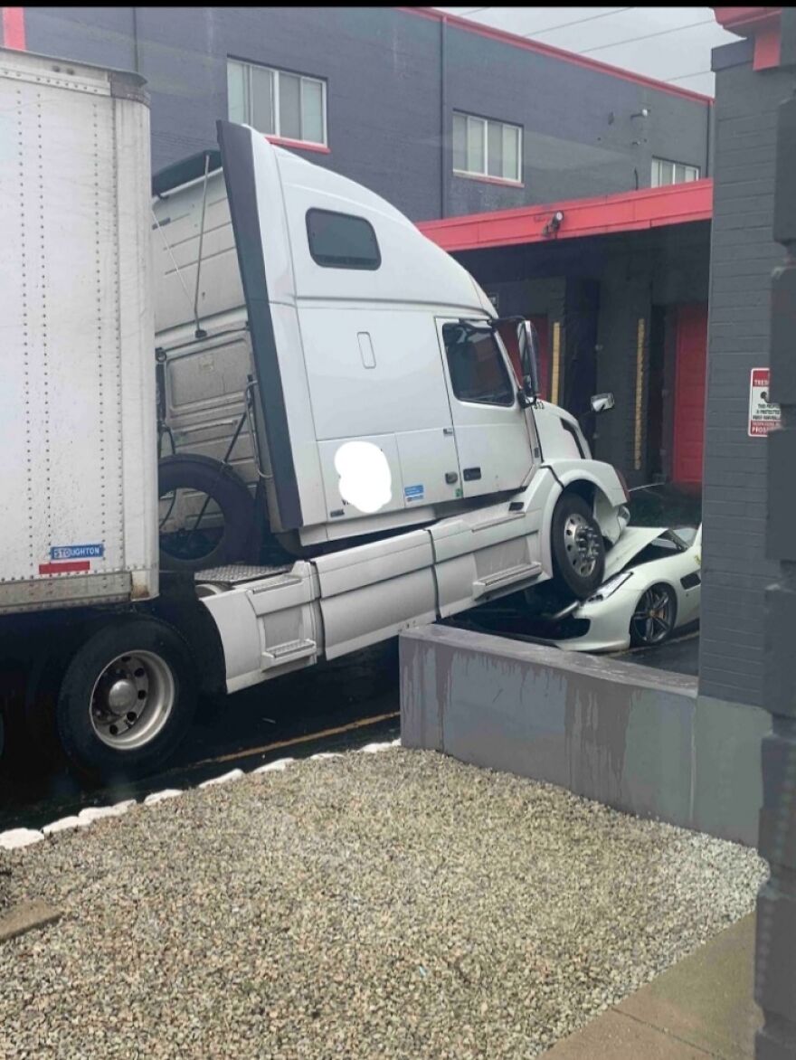 A semi-truck sits on top of a white sports car, causing an expensive accident and mistake in front of a building.