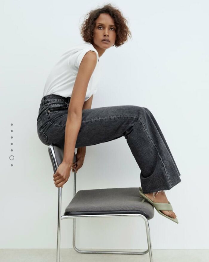 A woman in bizarre Zara content sitting backward on a chair, wearing a white t-shirt, dark jeans, and light green sandals.