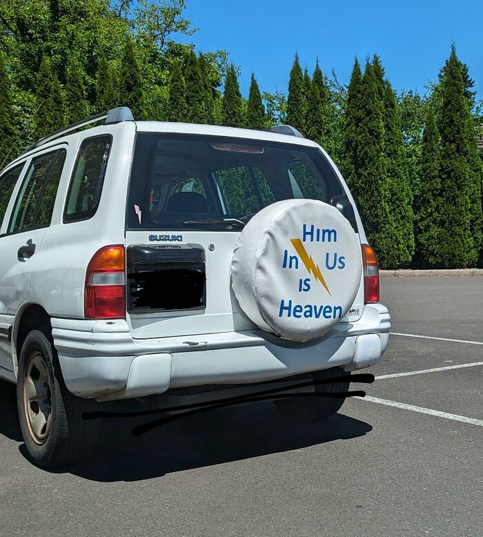 The back of a white Suzuki with a tire cover that reads Him in us is heaven, a confusing design fail.