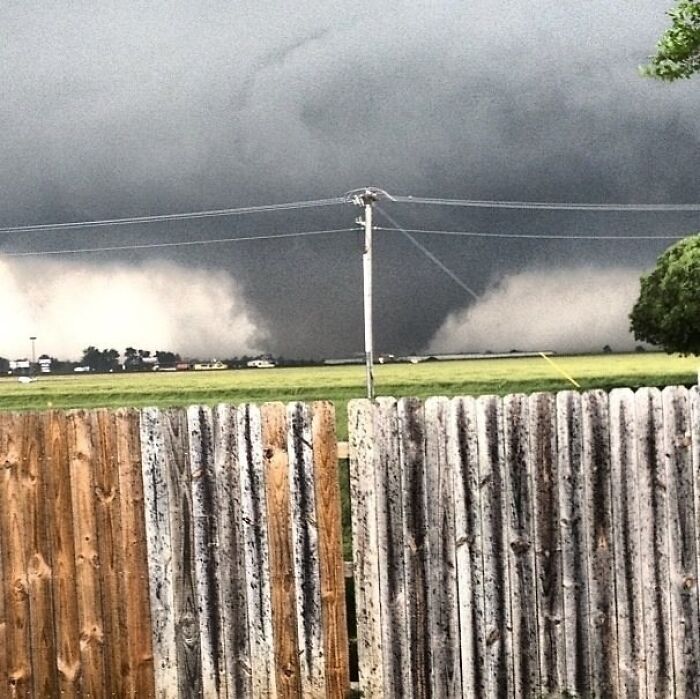 A large, dark tornado looms in the distance over a green field, seen from behind a wooden fence, illustrating different photos of the USA.