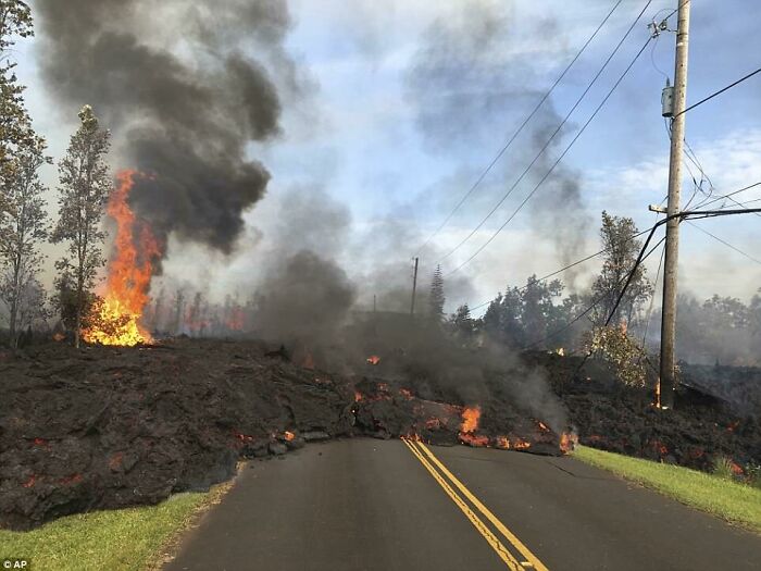 Molten lava flows across a road, with trees engulfed in flames and smoke rising, depicting fascinating photos of the USA's diverse landscapes.