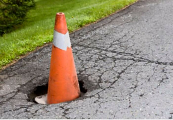 A vibrant orange traffic cone marking a prominent pothole on a cracked asphalt road in the USA.
