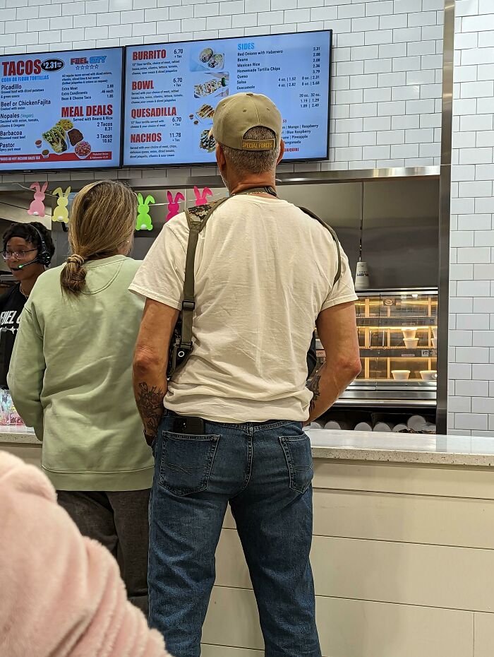 Man wearing a cap and holding a gun, standing in line at a fast-food restaurant in the USA.