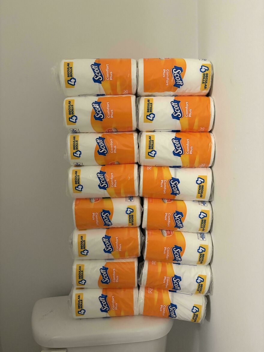 Many packs of Scott Comfort Plus toilet paper stacked high on a toilet tank, an amusing thing.